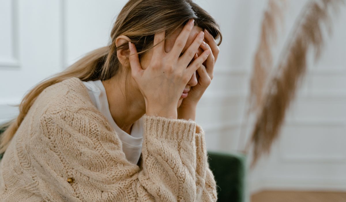 A person, possibly experiencing acute stress symptoms, sits with their hands covering their face, wearing a knitted sweater in a softly lit room with blurred decor in the background.