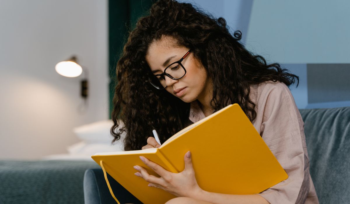 Person with curly hair and glasses writes in a large yellow notebook while seated indoors.