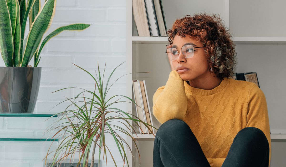A person in glasses sits indoors wearing a yellow sweater, leaning their head on their hand. They appear thoughtful. There are plants and shelves with books in the background.