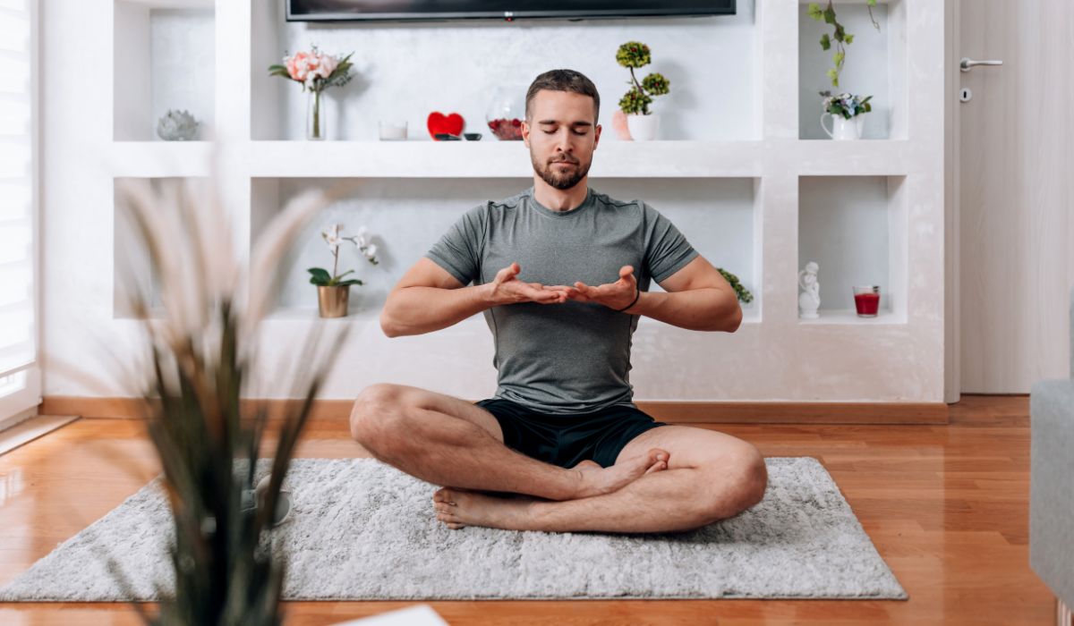 A man sits cross-legged on a rug in a living room, meditating with eyes closed and hands raised.