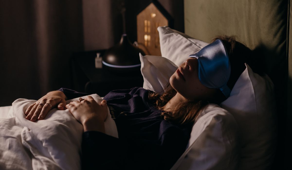 A person wearing a sleep mask lies in bed under white sheets, hands resting on the stomach; nearby, a nightstand with a lamp and diffuser quietly aids in managing intrusive thoughts for a peaceful night's rest.