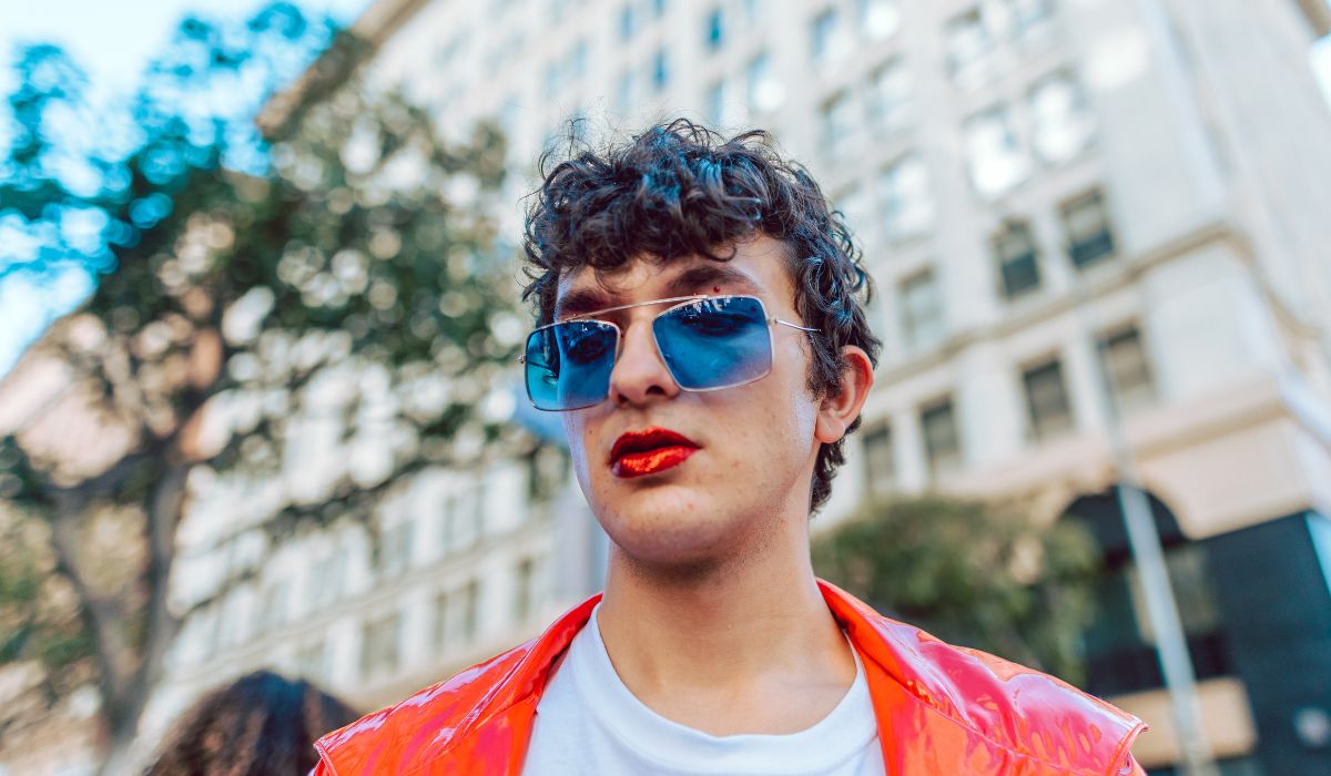 A person with curly hair wears blue sunglasses and red lipstick, dressed in a red jacket, standing in front of a building on a sunny day.
