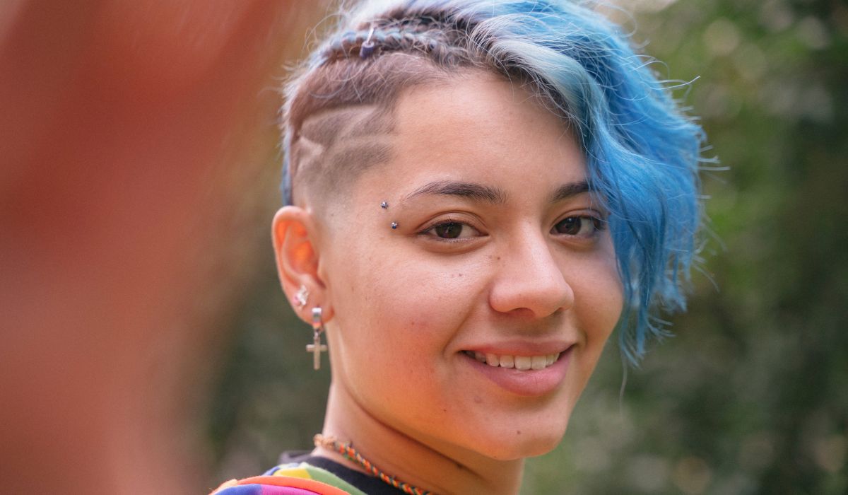 Person with short blue hair and an undercut, wearing earrings and a colorful shirt, smiling outdoors.
