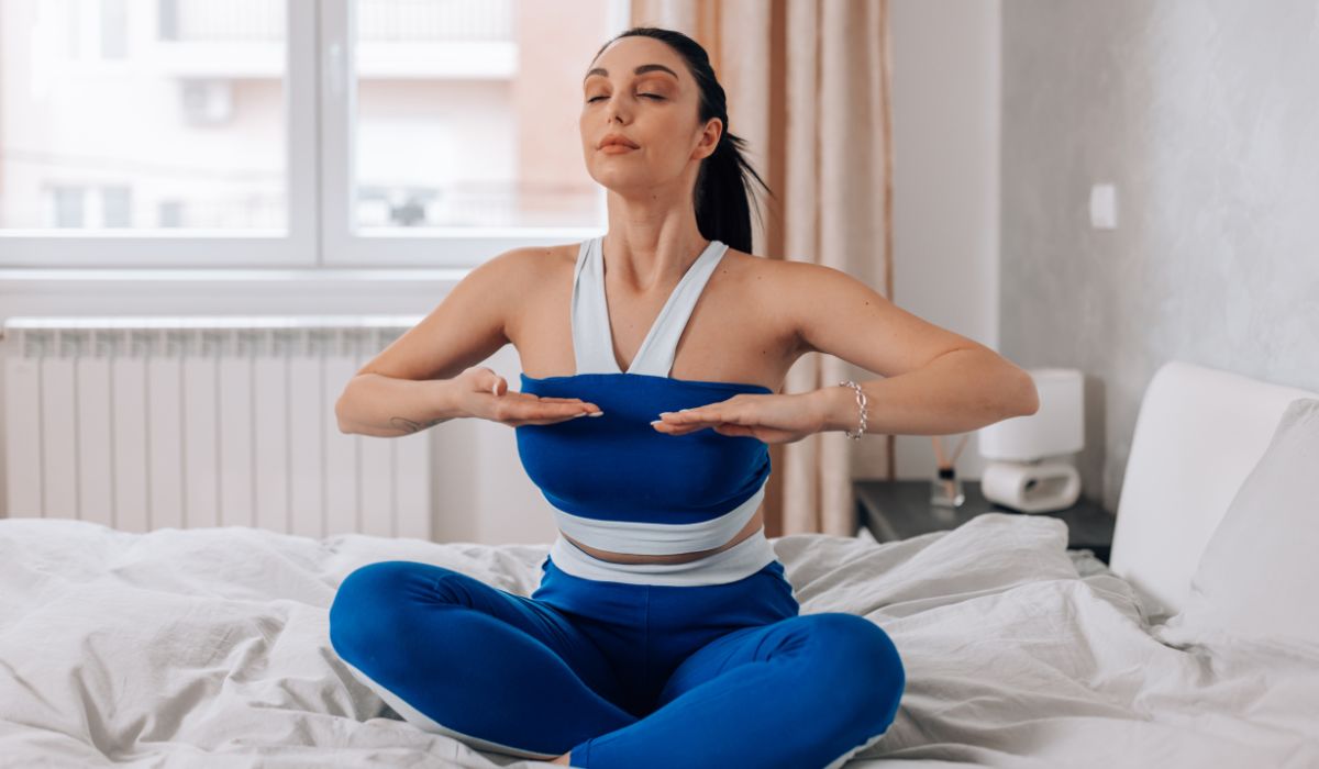 A woman in blue and white activewear sits cross-legged on a bed with eyes closed, holding her hands at chest height, appearing to practice deep breathing or meditation.