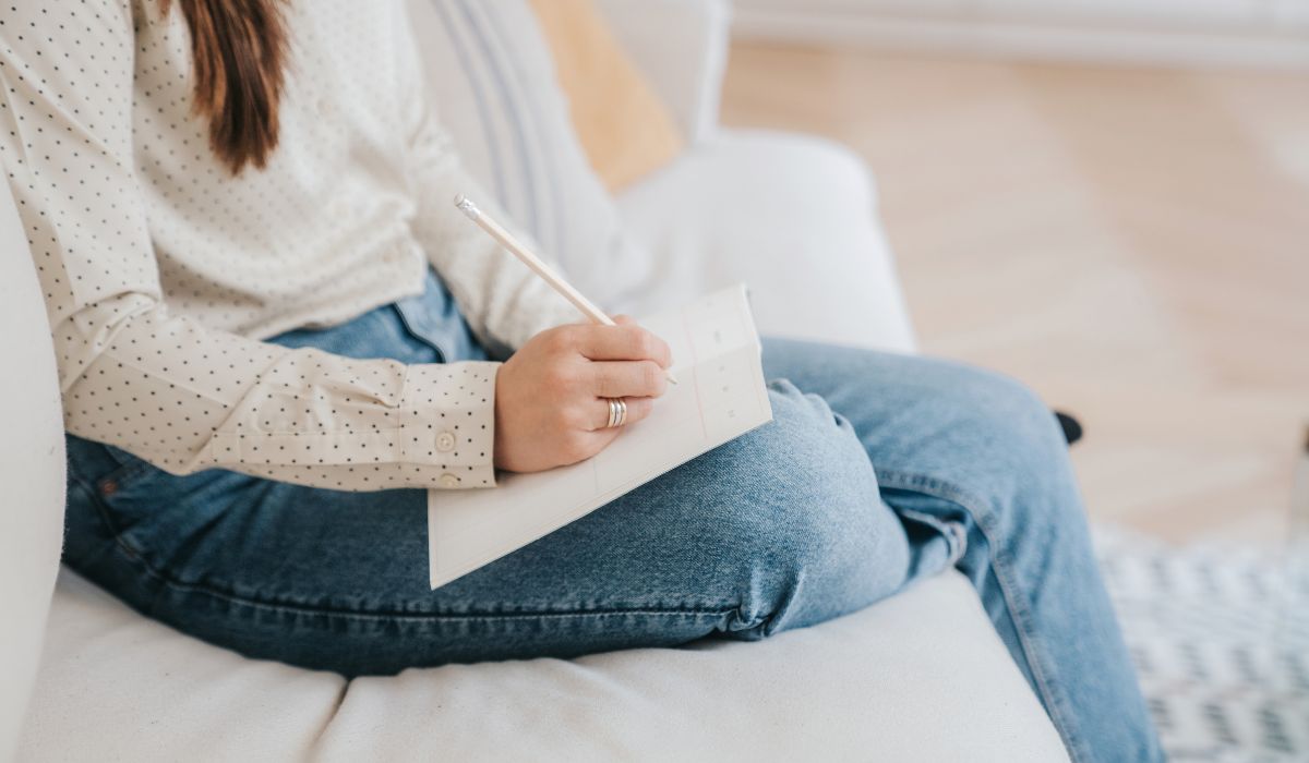 Person sitting on a couch, wearing a white polka dot shirt and blue jeans, writing in a notebook with a pen.
