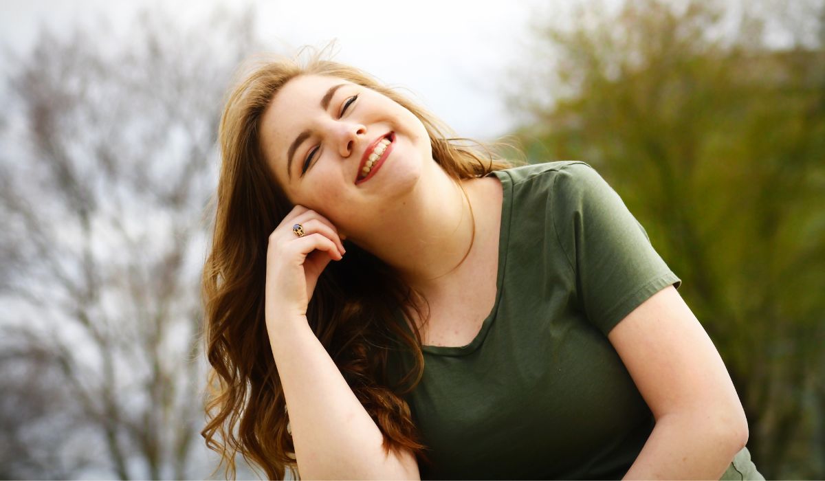 A woman with long brown hair wearing a green shirt smiles while sitting outdoors, with blurred trees in the background, perhaps reflecting on what causes perfectionism and how it affects daily life.