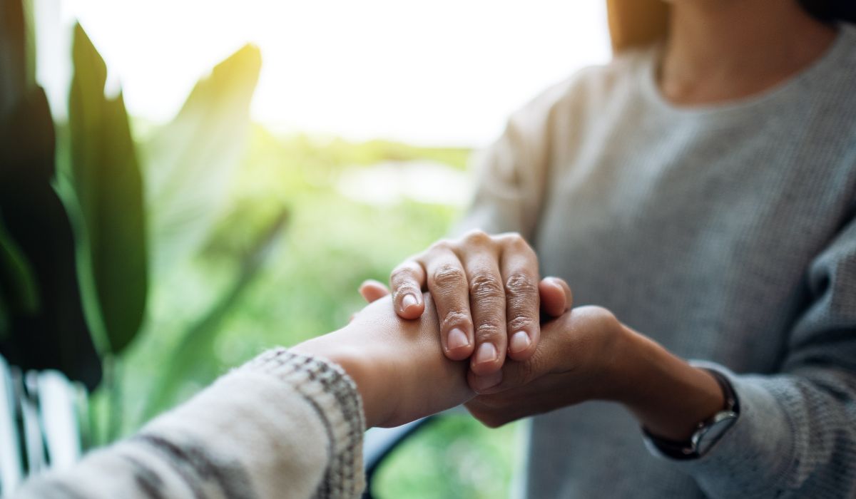 Two people are holding hands, with one person gently placing their hand over the other's—a comforting gesture often used in anxiety interventions, indoors with greenery visible in the background.