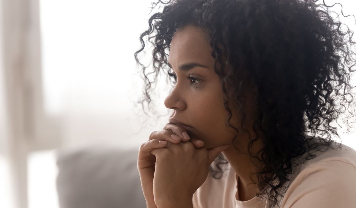 A woman with curly hair sits indoors, resting her chin on her clasped hands, looking thoughtful and concerned, as if contemplating the results of a trauma quiz.
