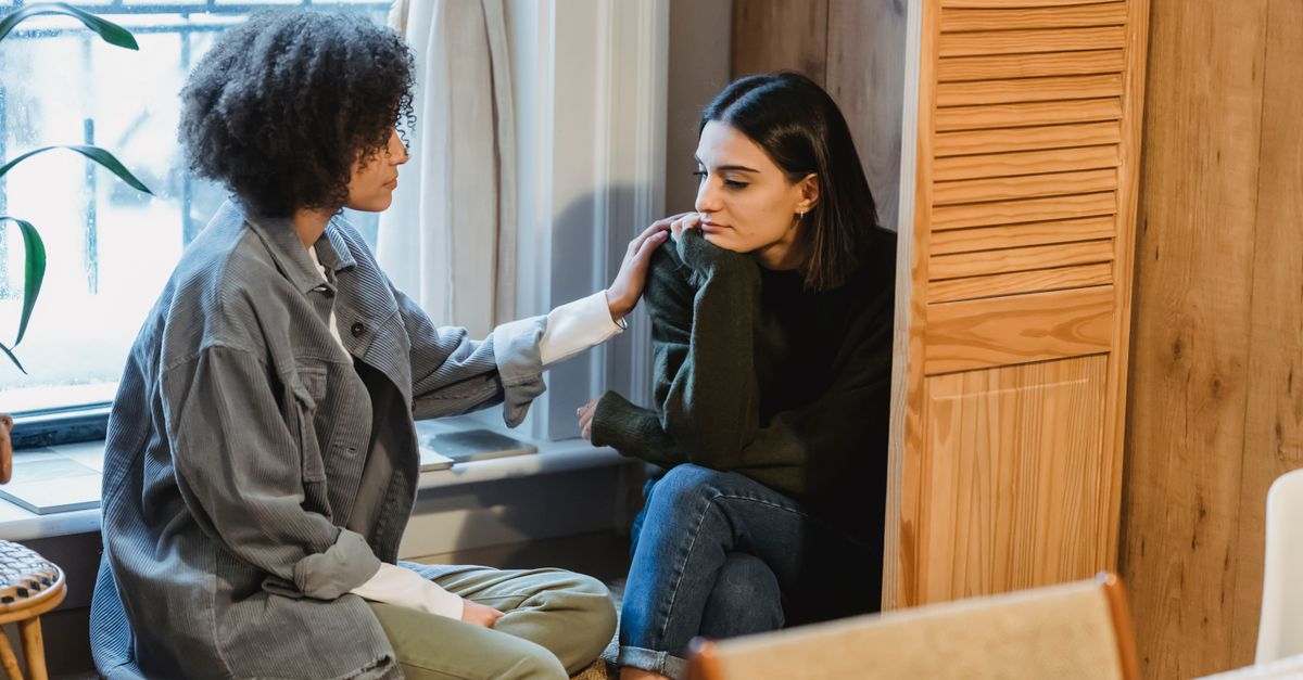 Calm young multiracial female best friends in casual clothes sitting near window and home and discussing discussing problems