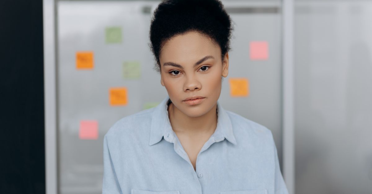 A woman wearing a blue shirt poses confidently in an office with sticky notes on the glass wall behind her.