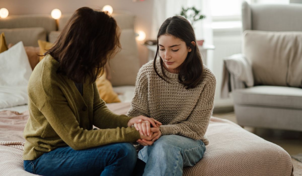 Two women sit on a bed facing each other, holding hands and having a serious conversation in a softly lit bedroom, discussing how to come out to your parents.