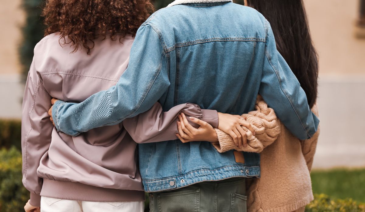 Three people standing outdoors with their backs to the camera, arms around each other, wearing casual jackets and sweaters.