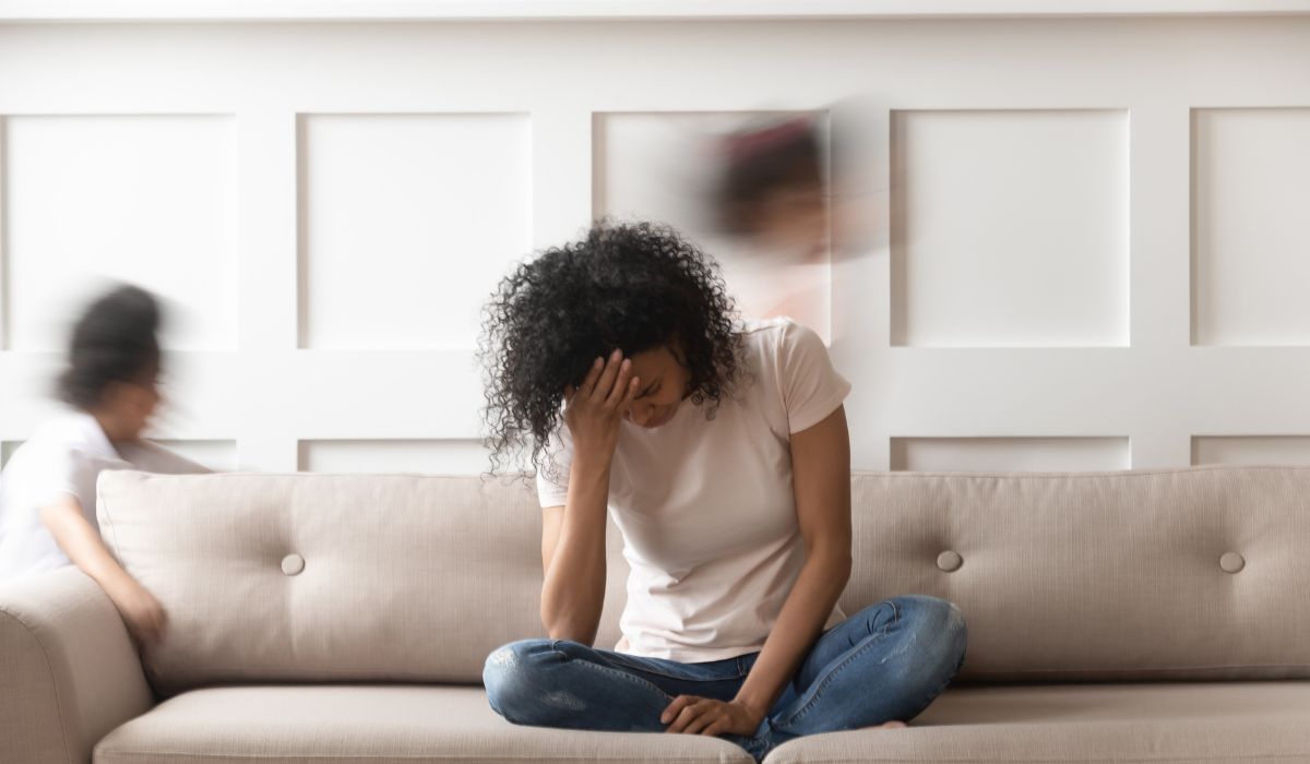 A woman sits on a couch with her head in her hand, appearing stressed, while two children move quickly in the background—signs she may be outside her window of tolerance.
