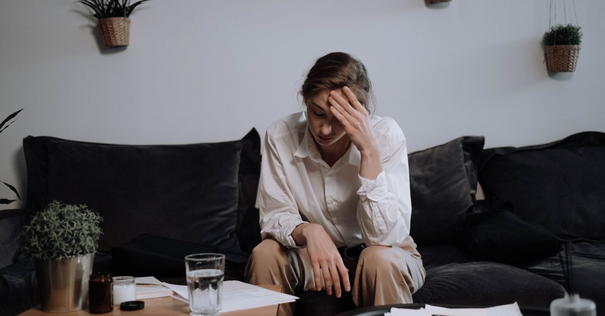 Woman experiencing stress and headache while sitting on a sofa at home, surrounded by papers.