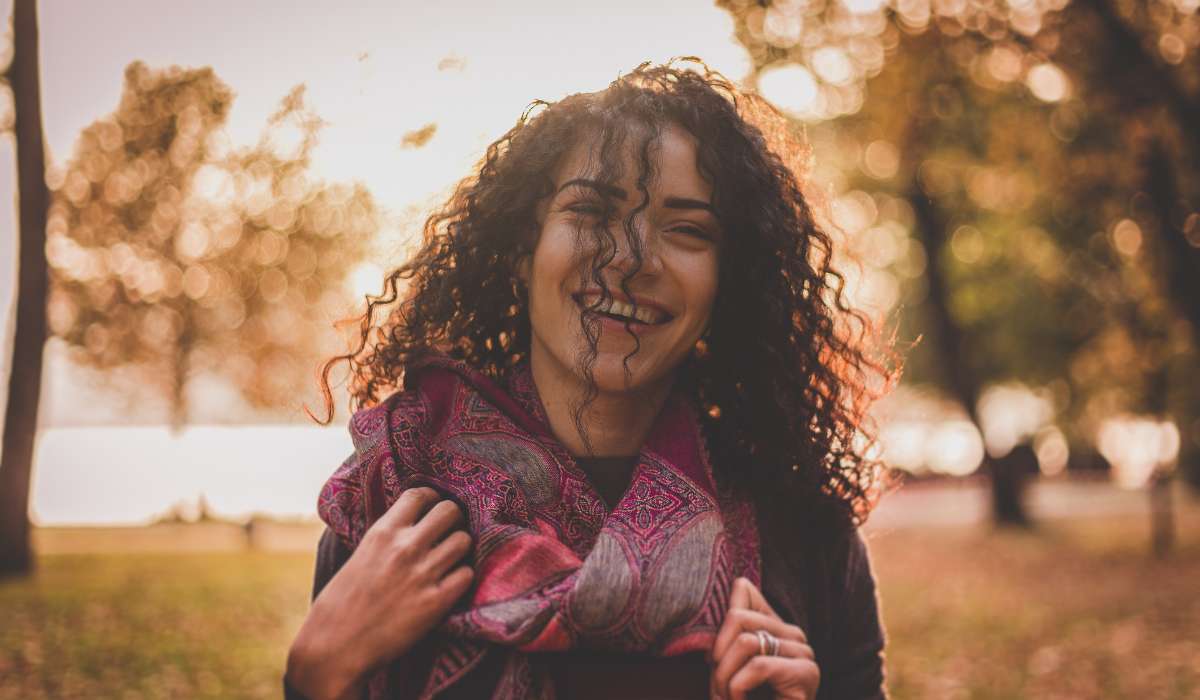 A person with curly hair smiles outdoors while holding a patterned scarf, with autumn trees blurred in the background.