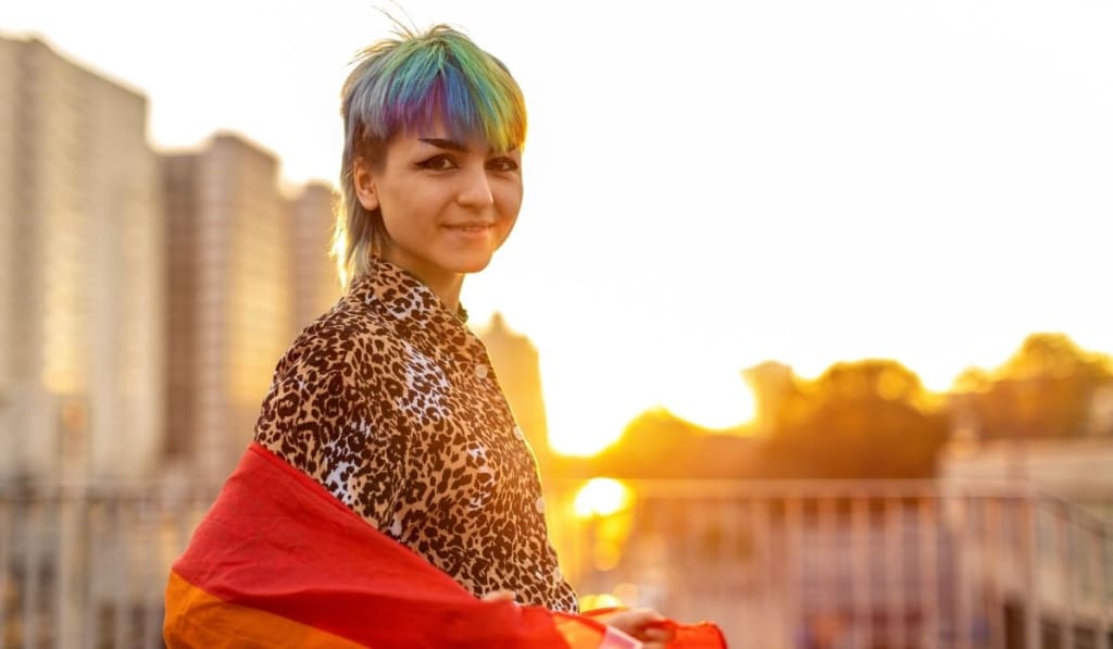 A person with short, multicolored hair and a leopard print shirt stands outdoors at sunset, draped in a rainbow flag—showing that coming out is important and celebrating identity with pride.