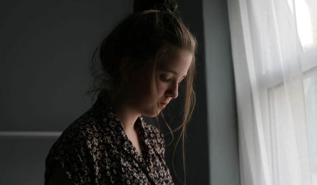 A young woman with long hair looks down thoughtfully near a window with sheer curtains, illuminated by soft natural light, perhaps reflecting on her experiences with EMDR side effects.
