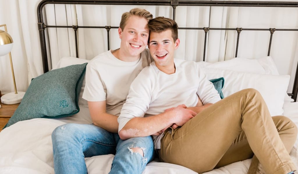 Two young, gender diverse men sit closely together on a bed, smiling at the camera. One wears a white shirt and jeans, the other a white shirt and tan pants.