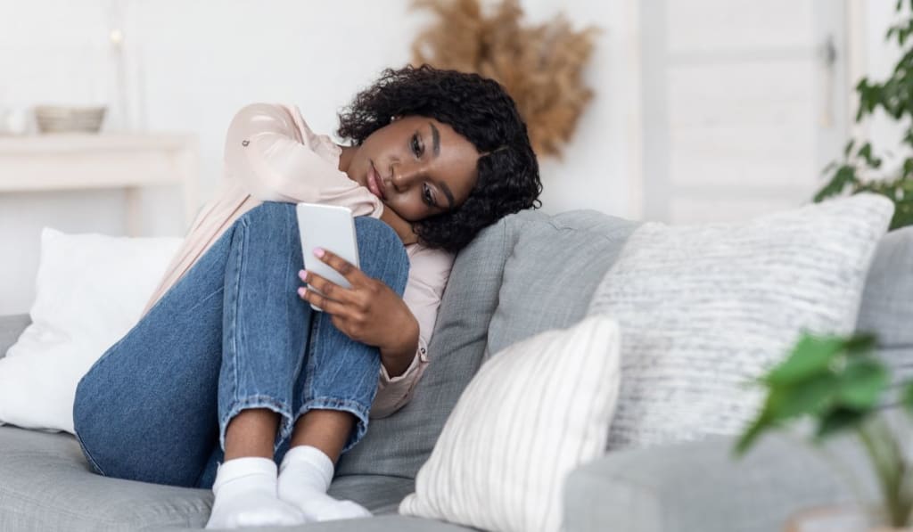A woman sits on a couch, looking at her phone with a neutral expression, as if lost in thought. Resting her head on her knee, she seems to drift toward a sense of self-abandonment.