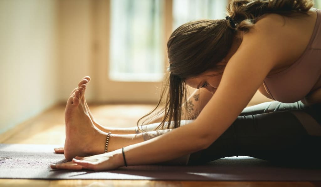 Person in a yoga pose, seated forward fold, on a mat indoors with sunlight streaming through a window.