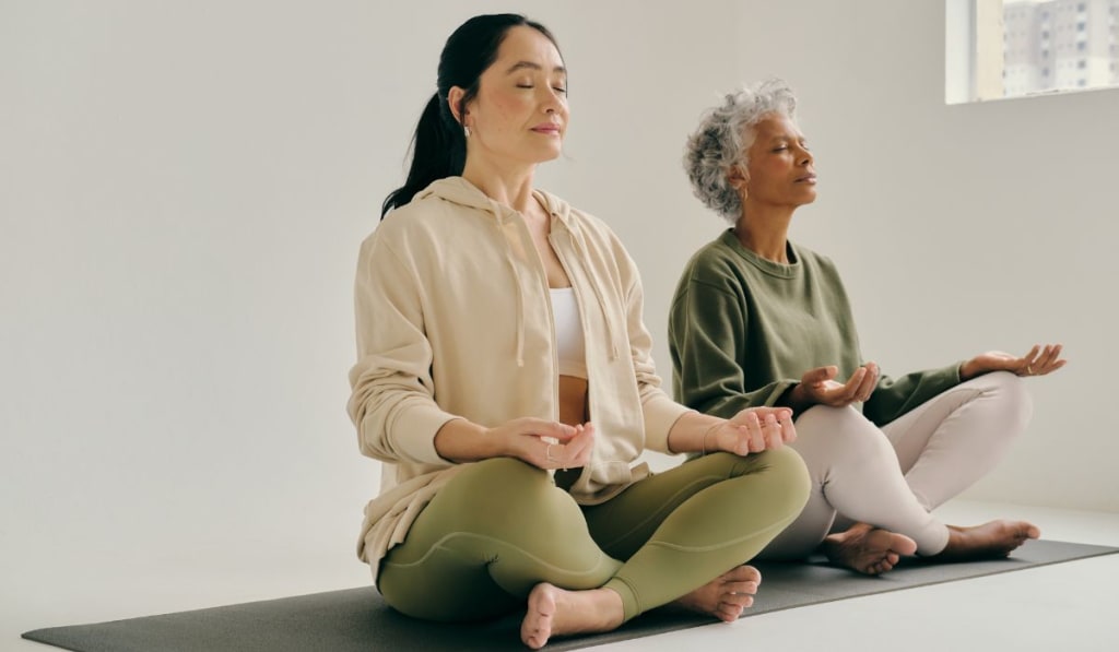 Two people sit cross-legged on mats in a bright room, meditating with eyes closed, finding tranquility amidst life's acute stress symptoms.