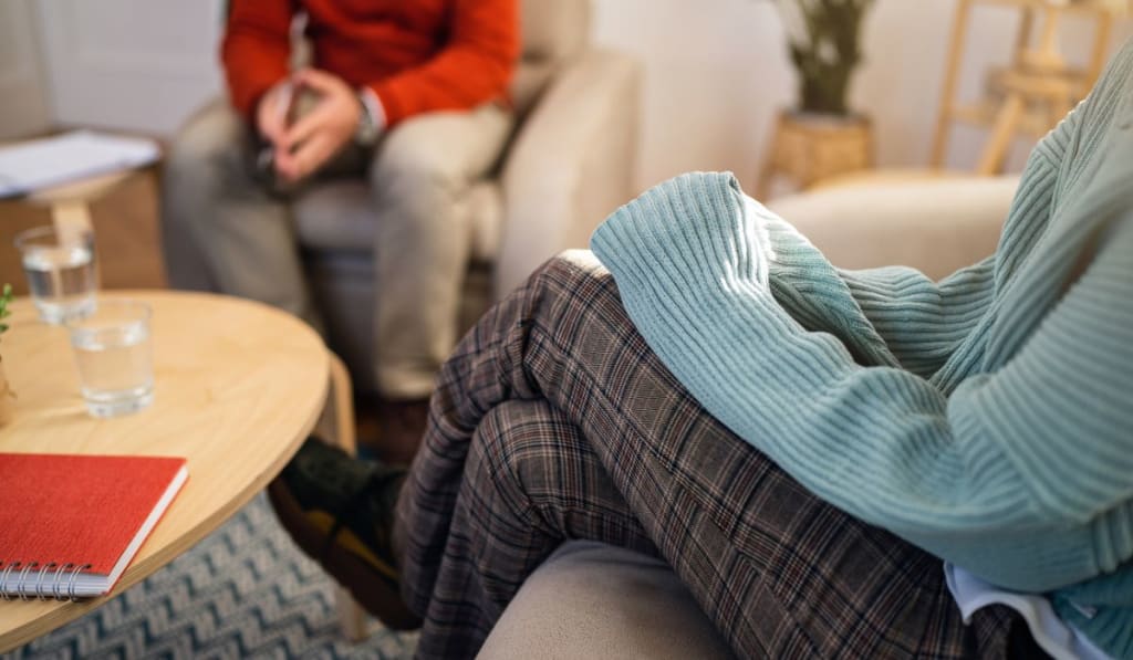 Two people are seated in a room, one wearing a turquoise sweater and plaid pants, the other in red. Nearby, a wooden table holds a notebook and glasses of water as they discuss the difference between acute stress and PTSD.