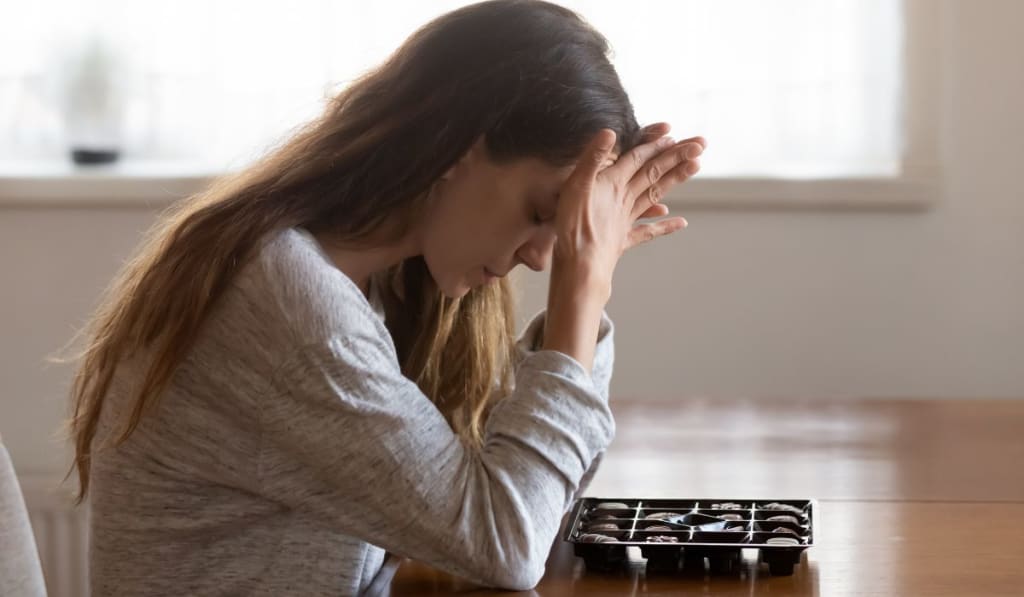 A woman sits at a table with her head in her hands next to an open box of chocolates, appearing stressed or upset—an image that captures the emotional weight perfectionism can bring.
