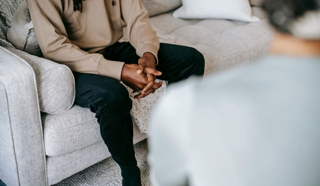 A person sits on a light-colored couch with hands clasped, facing another individual whose back is partially visible in the foreground, suggesting a thoughtful moment—perhaps during a trauma quiz or discussion.