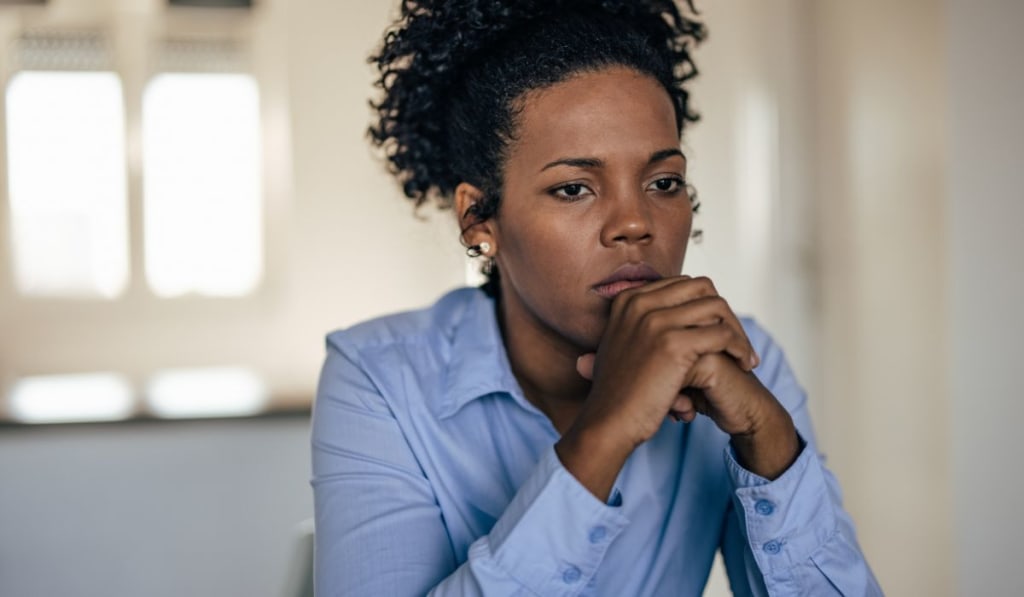 A person with curly hair and a blue shirt sits indoors, looking pensive with their hands clasped near their chin—a subtle display of social hypervigilance.