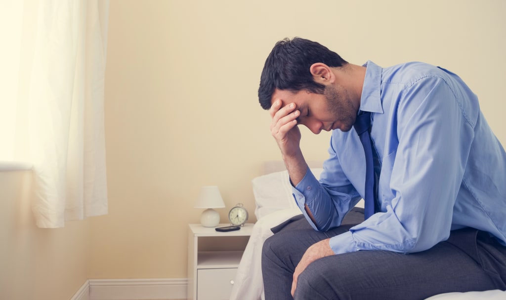 A man in a blue shirt and tie sits on a bed with his head in his hand, visibly stressed—a moment of reflection on the difference between burnout vs. depression. A man in a blue shirt and tie sits on a bed with his head in his hand, visibly stressed—a moment of reflection on the difference between burnout vs. depression.