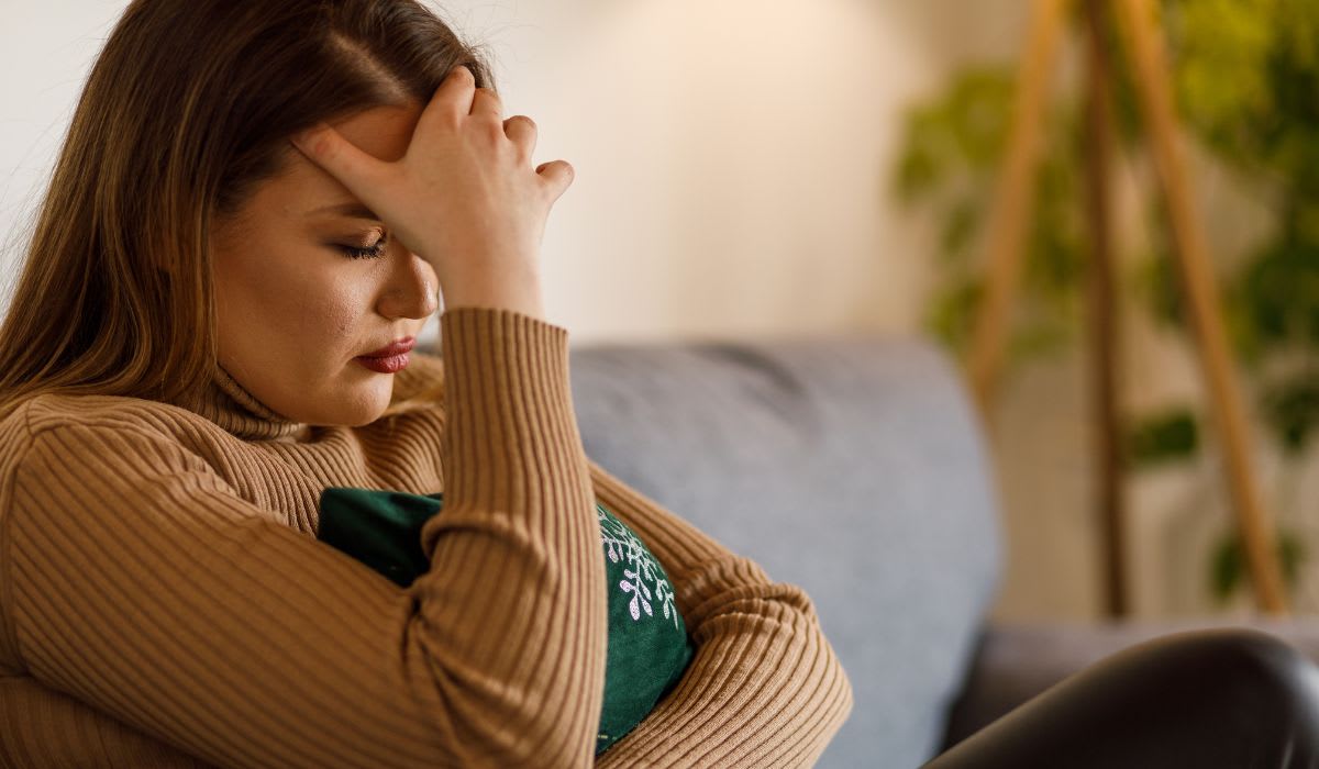 A woman sitting on a couch holds her forehead with one hand and rests the other on a pillow.