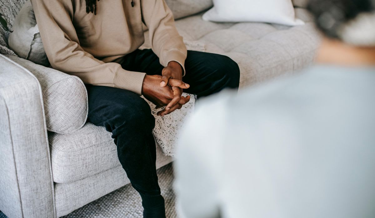 Person sitting on a couch with hands clasped, facing another individual whose back is visible in the foreground—capturing a reflective moment often explored in an emotional trauma test.