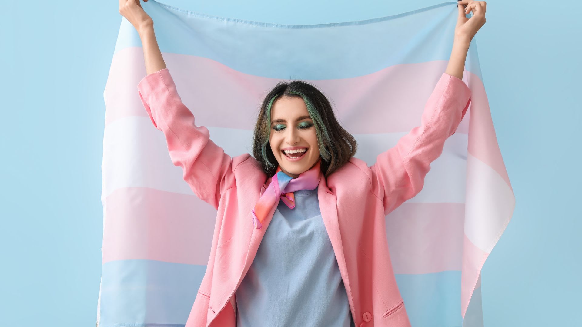 A person with long hair and colorful makeup smiles with eyes closed, holding a blue, pink, and white striped flag above their head while wearing a pink blazer and colorful neck scarf.