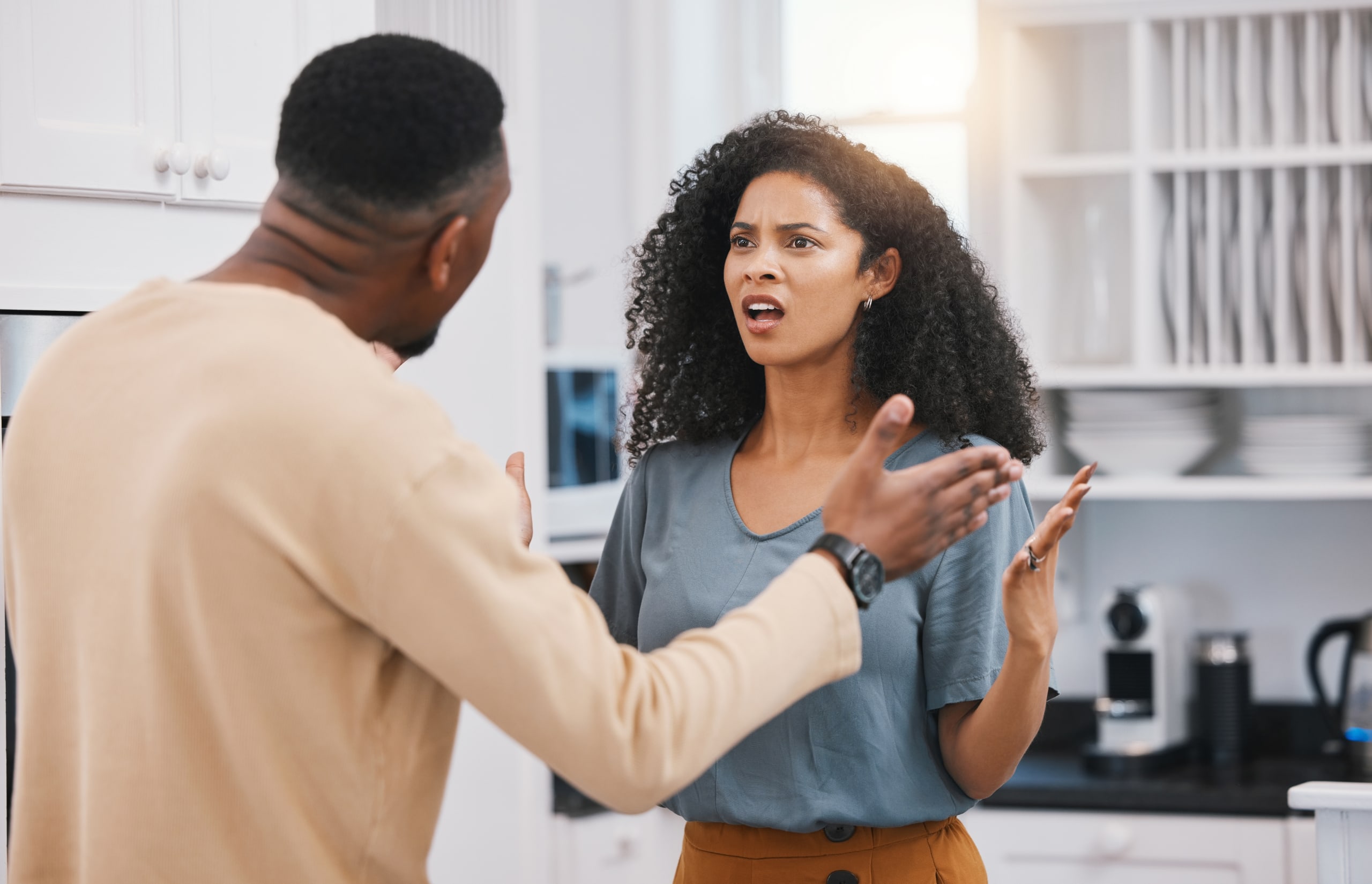 A man and woman are standing in a kitchen having an animated discussion. The woman appears surprised or confused.
