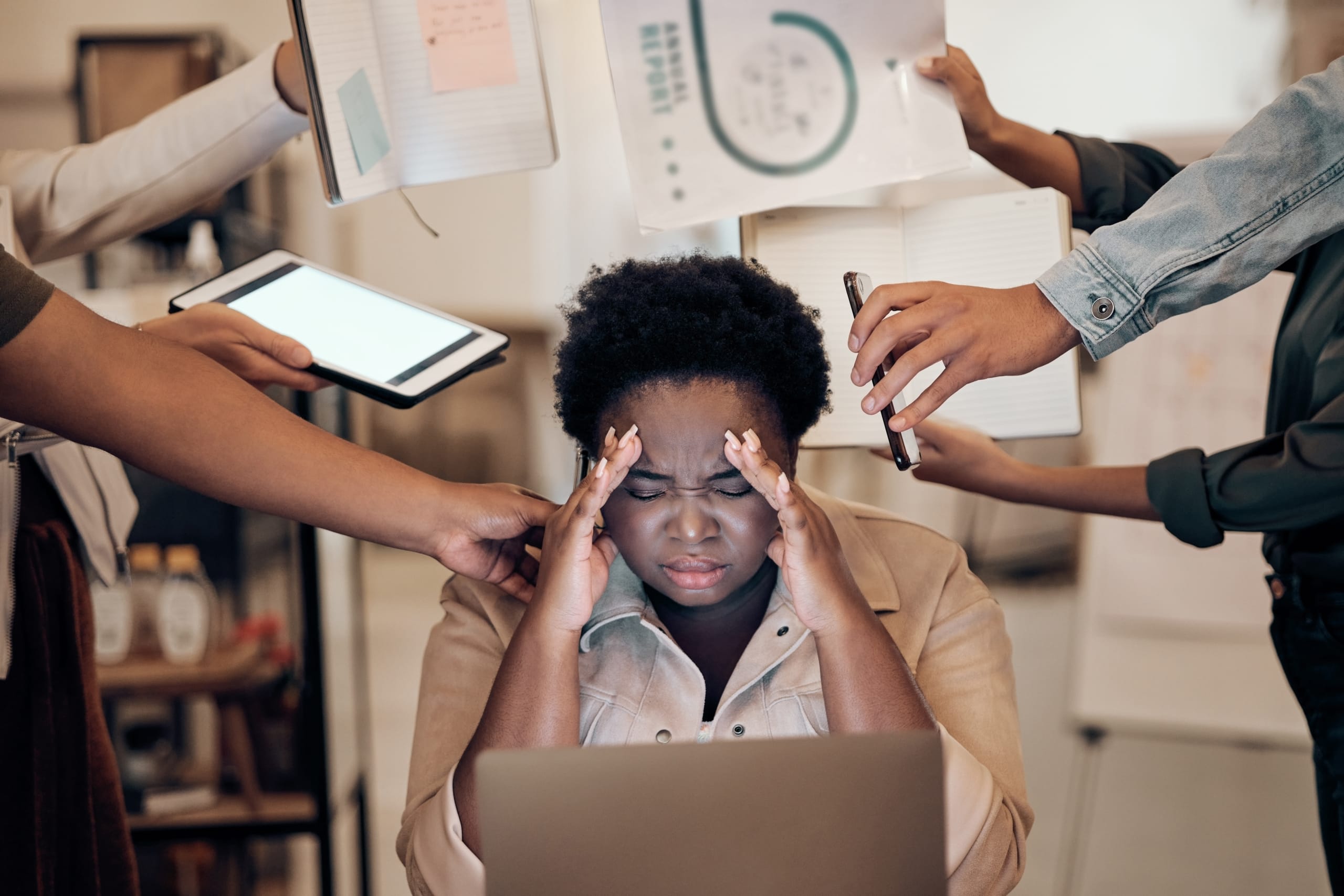 A woman sits at a desk, appearing stressed, with her hands on her temples. Several hands around her hold a tablet, phone, and documents, indicating a busy and demanding environment.
