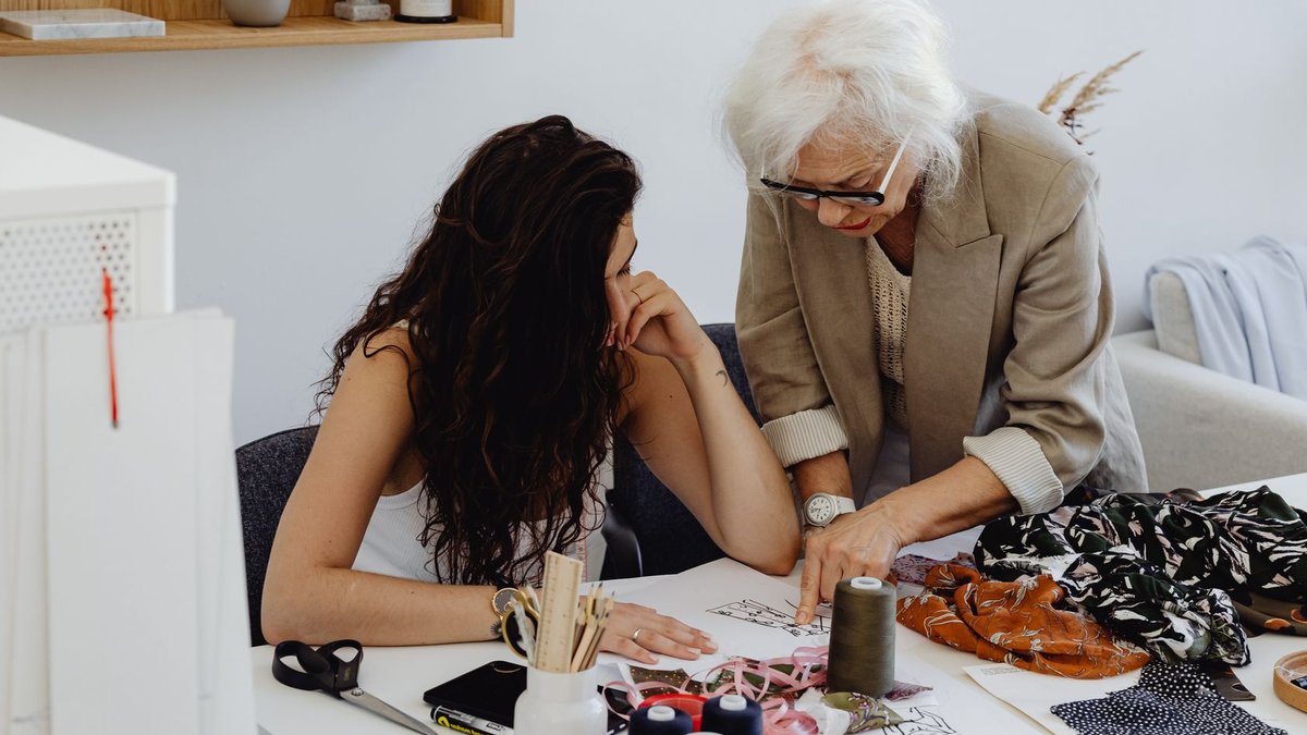 Designers Use AI to Sell Custom Collections Online Elderly woman instructs young designer at worktable, sharing expertise in a fashion studio.