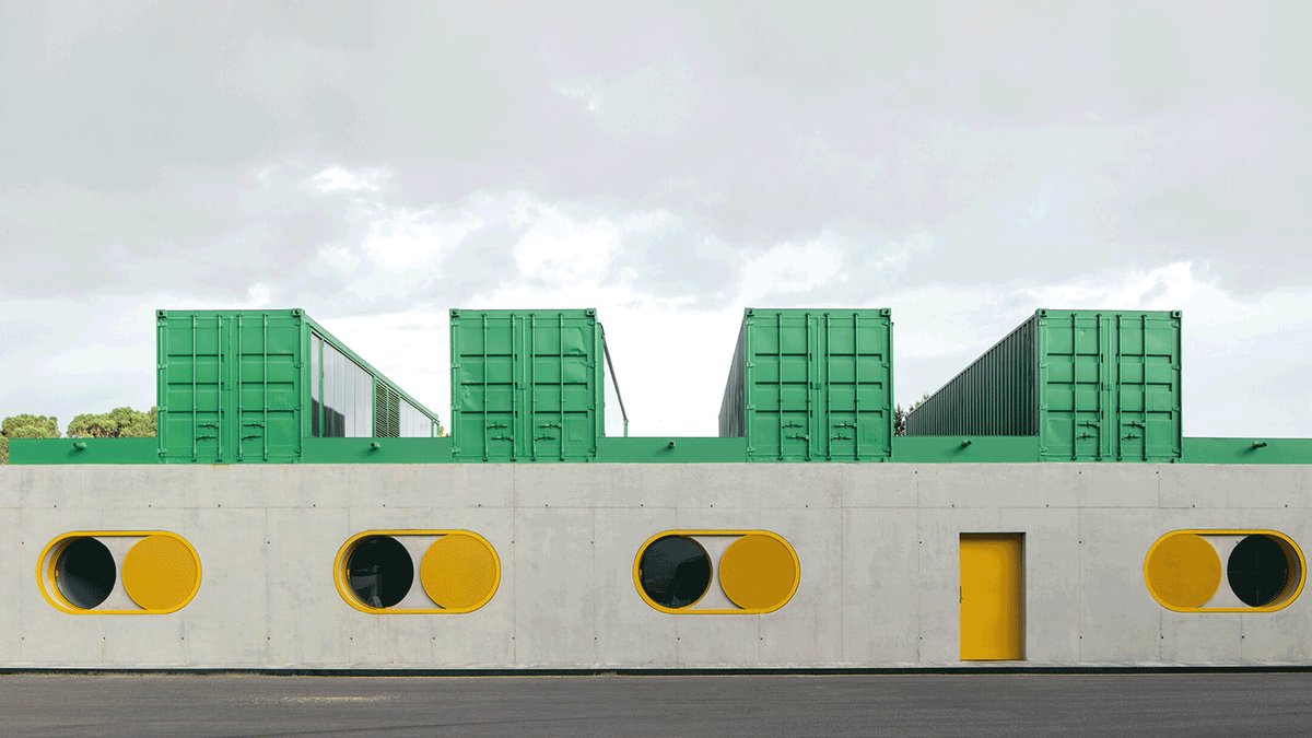A modern building with green shipping containers on the roof, gray concrete walls, a yellow door, and circular yellow-framed 