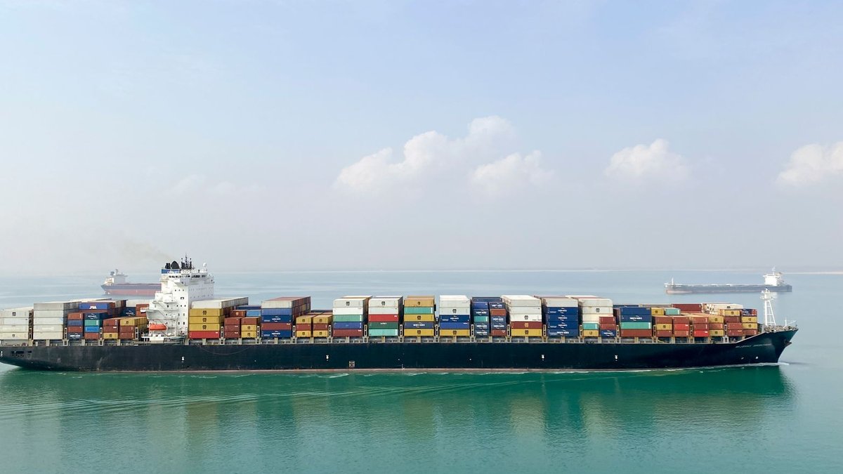 Shipping Rates Surge Amid Declining Cargo Volumes Aerial view of a large cargo ship filled with containers sailing on a calm sea under a clear sky.