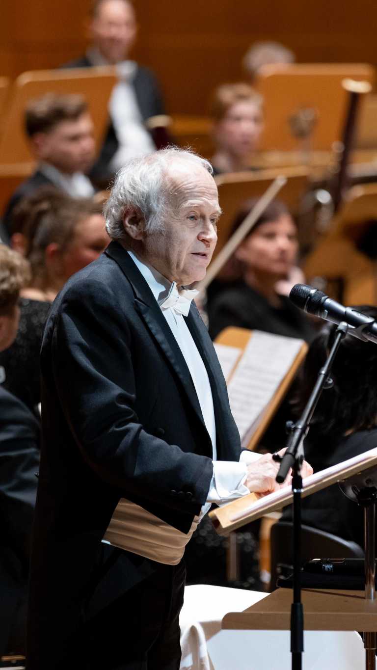 Dirigent Adam Fischer hält auf einer Bühne eine Rede vor einem Mikrofon