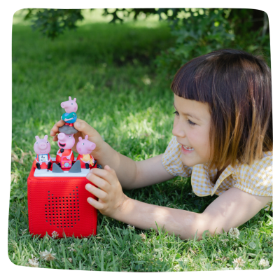 Young female toddler playing in the grass with her red Toniebox and several Peppa Pig Tonies.