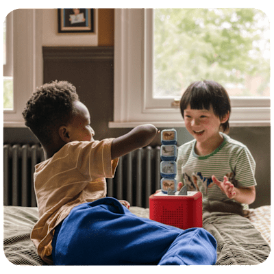 Two young boys, one black child with short cropped hair and one asian child with a bowl cut, playing on a bed together with a red Toniebox and four stackable clever Tonies.