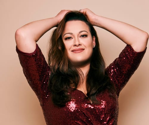 Ashley Brown with her hands on her head against a light orange backdrop