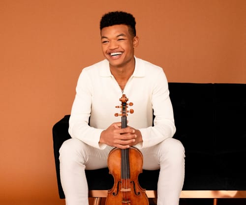 Randall Goosby sitting with his violin against an orange wall