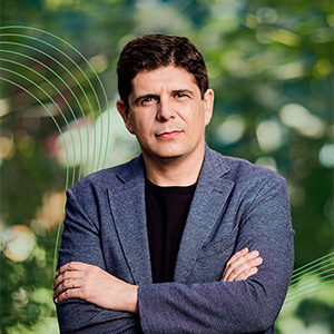 Javier Perianes wearing a grey suit jacket and black shirt with his arms crossed in front of blurred garden backdrop.