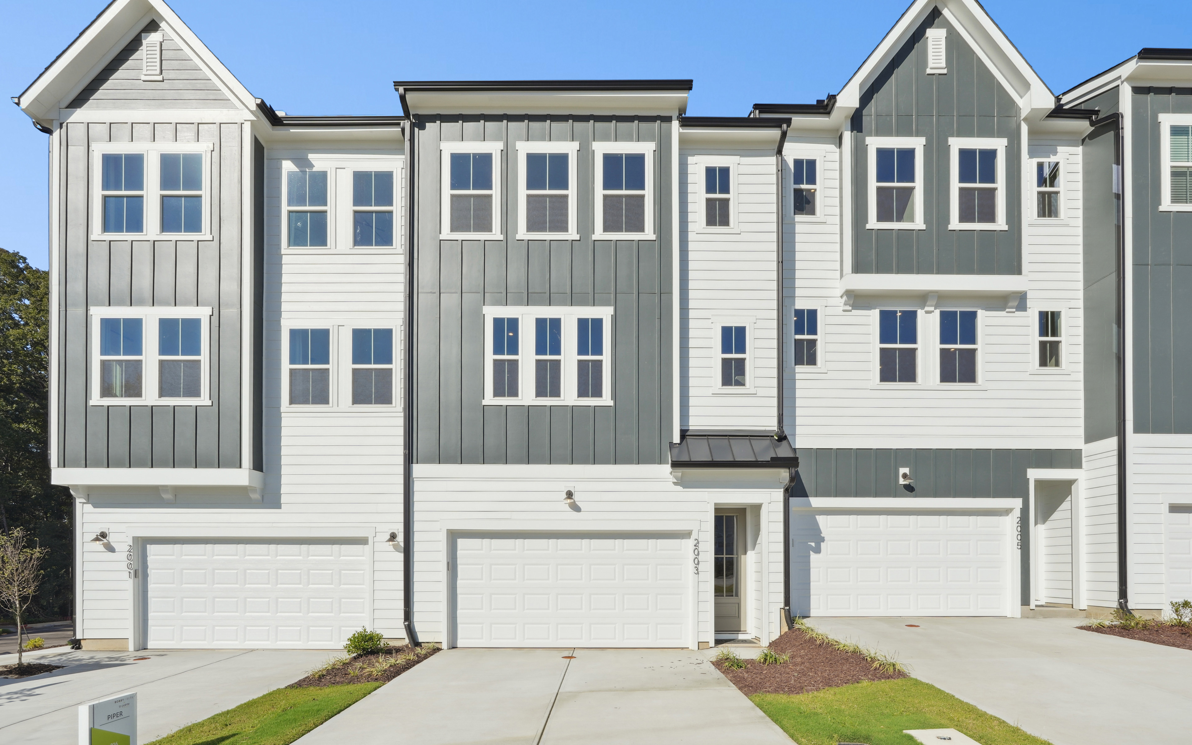 A row of modern, multi-story townhouses with gray and white siding, gabled roofs, and attached garages, set against a clear blue sky.