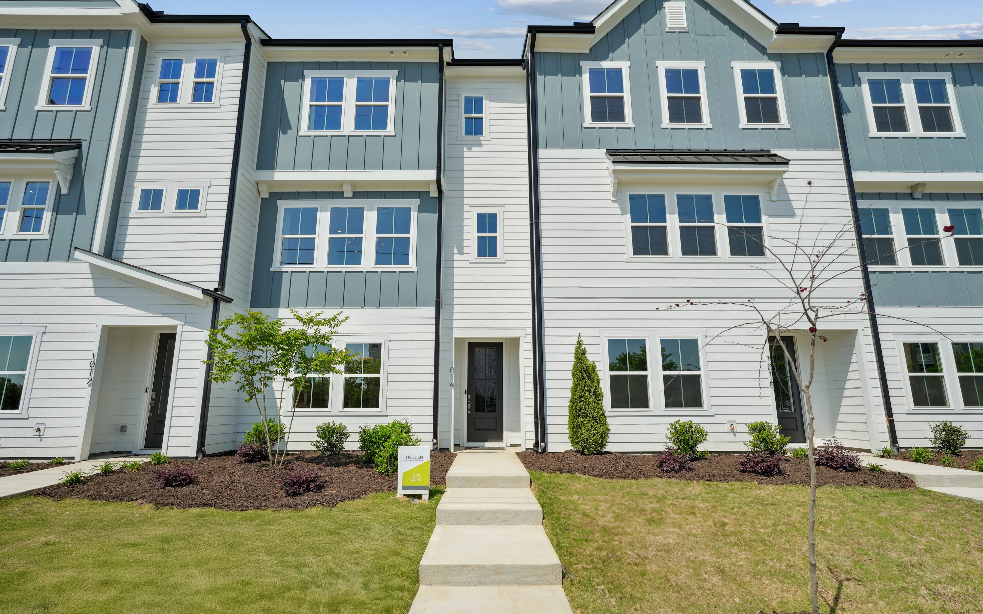 A multi-story townhouse community with a well-manicured lawn and landscaping in the foreground, featuring a mix of siding colors and architectural styles.