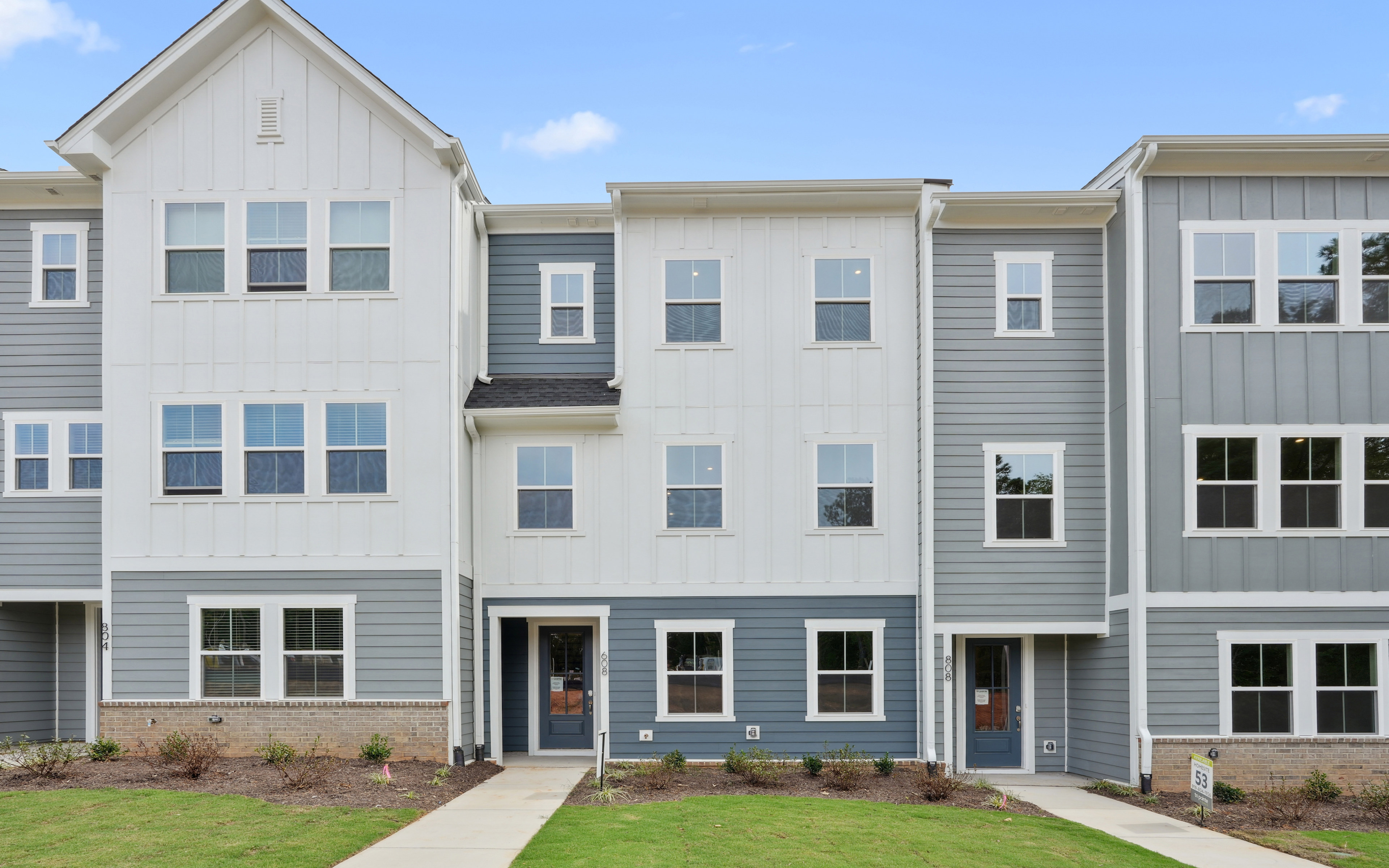 A multi-story townhouse complex with gray siding, white trim, and a grassy lawn in the foreground.