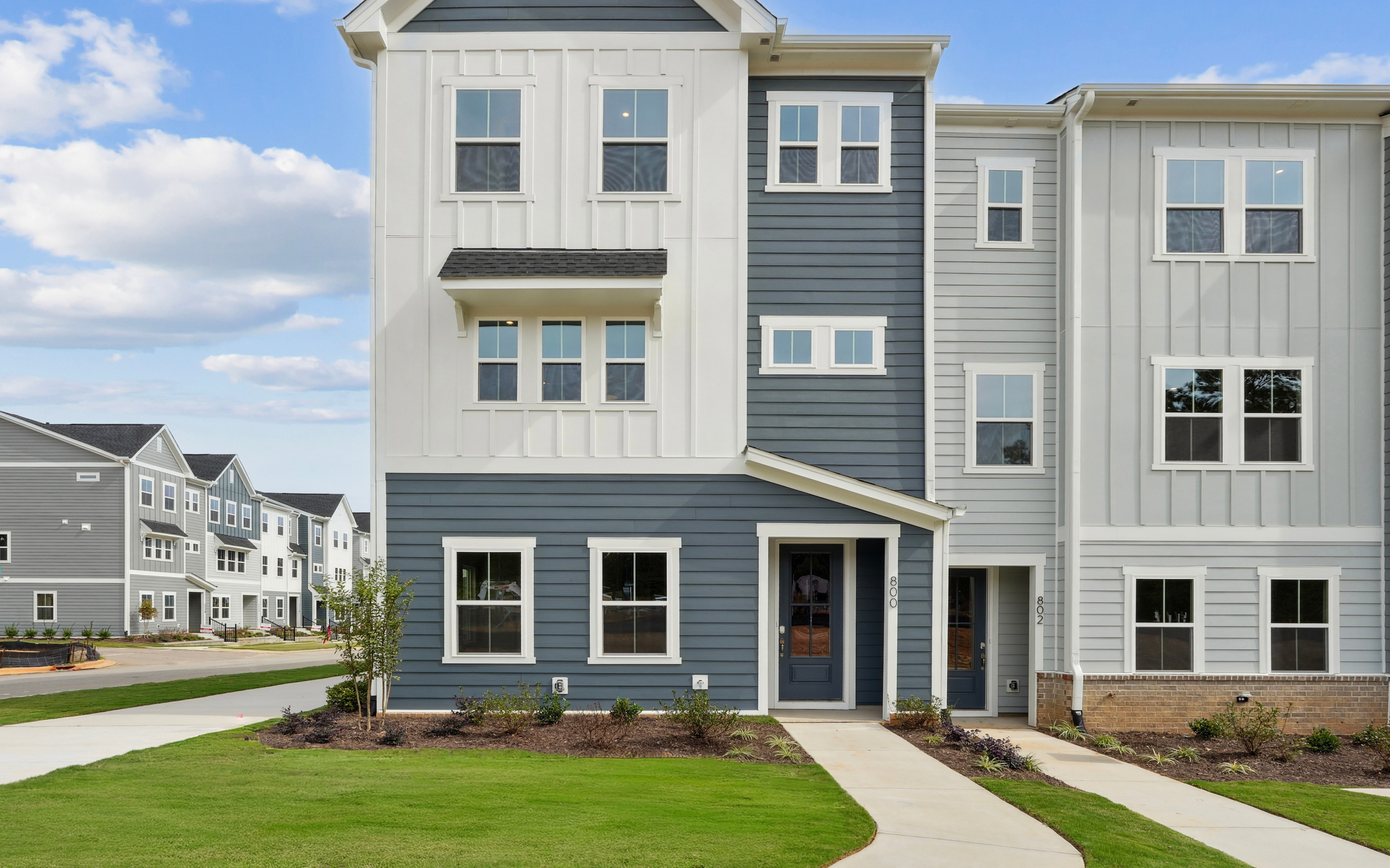 A multi-story residential building with a gray exterior and white trim, surrounded by a well-manicured lawn and set against a blue sky with scattered clouds.