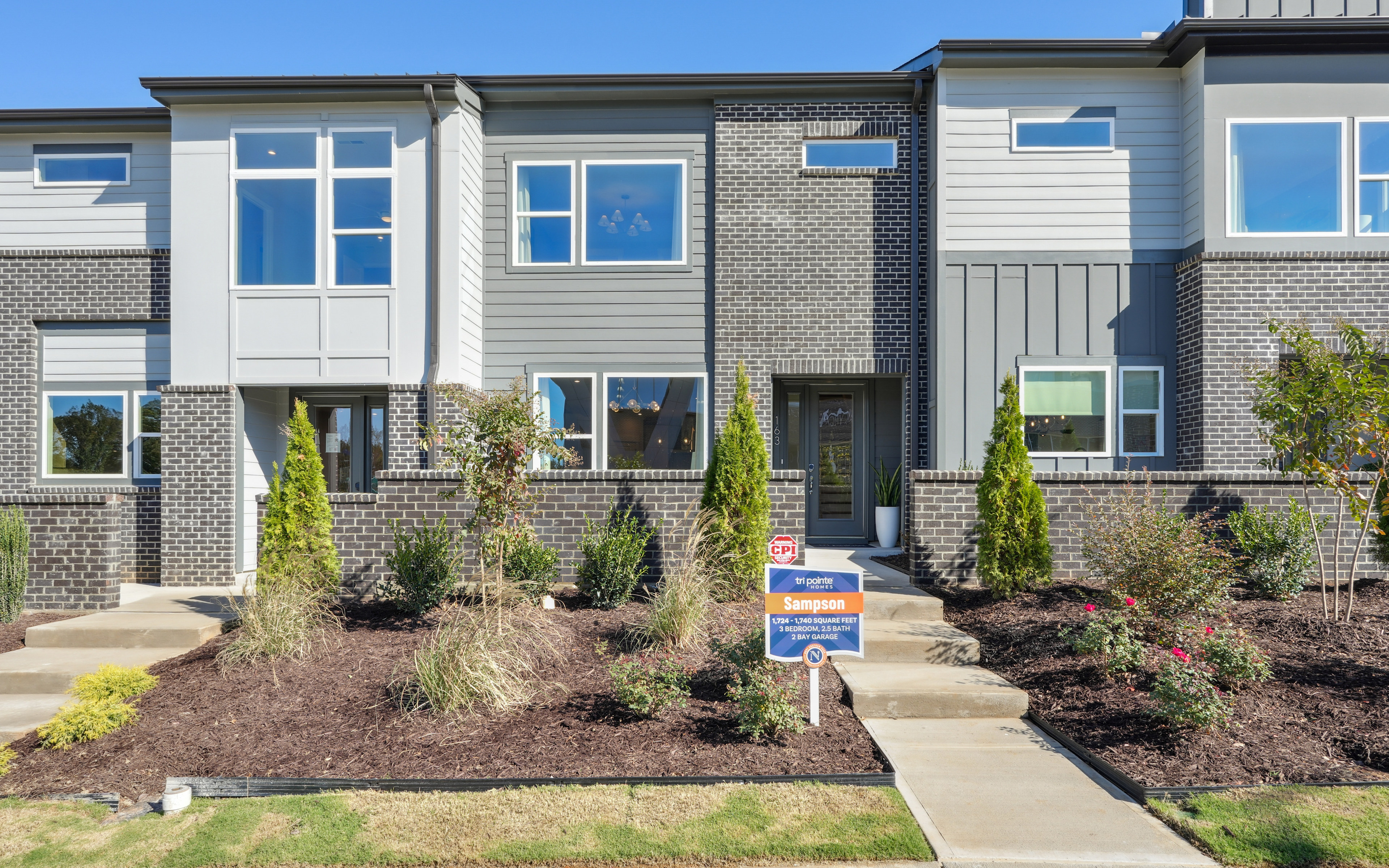 A modern townhouse with a well-landscaped front yard, featuring a brick and siding exterior, large windows, and a paved walkway leading to the entrance.