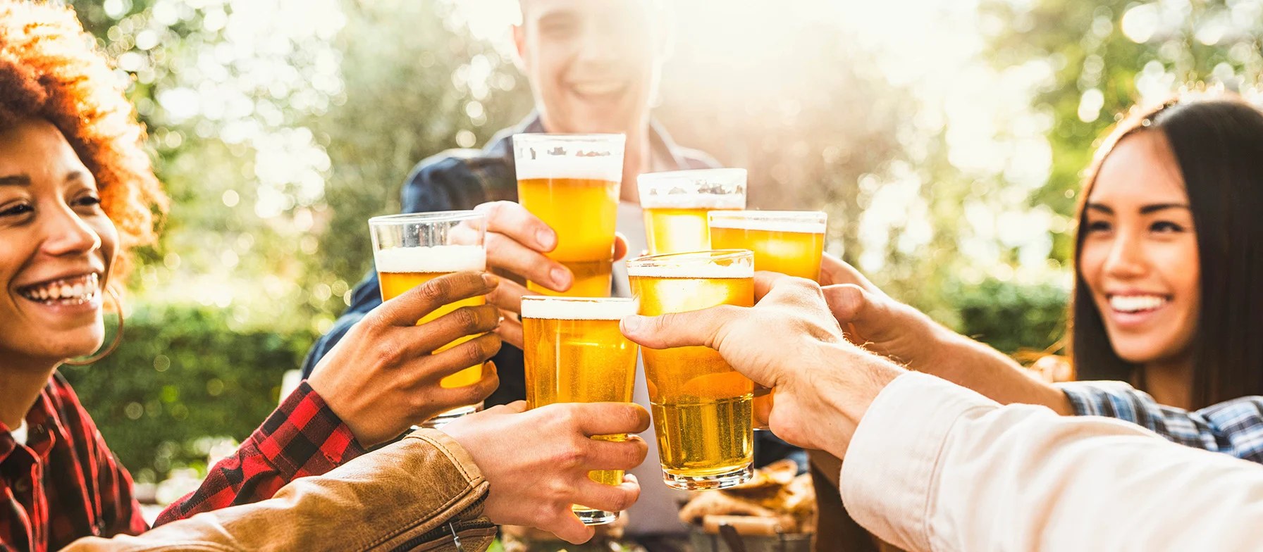 A group of friends are gathered outdoors, raising their glasses of beer in a celebratory toast, with a lush, green background providing a pleasant setting for their gathering.